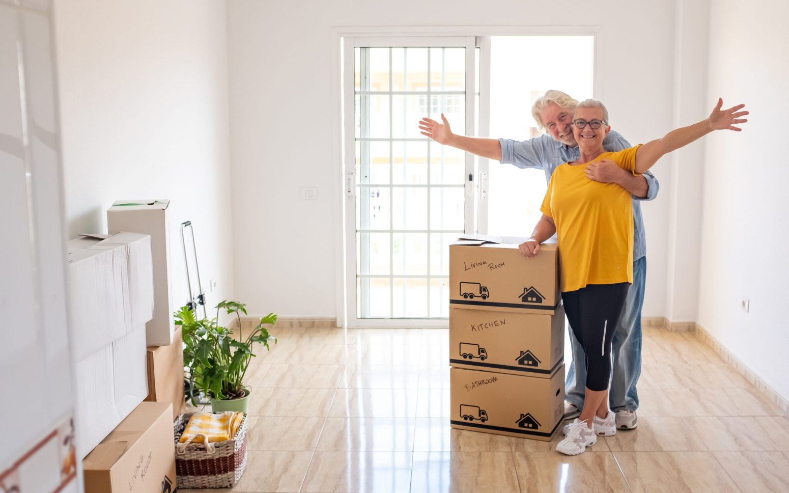 Smiling caucasian senior couple white haired open arms looking at camera during relocation, happy for the new beginning. Moving boxes on the floor, empty apartment